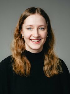 Frau mit langem braunem Haar lächelt in die Kamera, trägt schwarzen Rollkragenpullover.