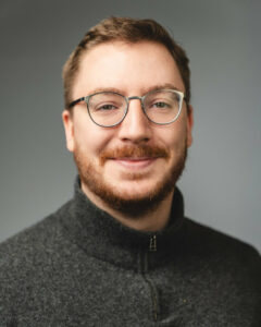 Mann mit Brille, Bart und grauem Pullover vor grauem Hintergrund.