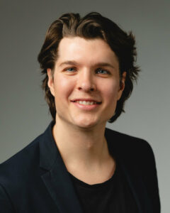 Mann mit dunklem Haar und Sakko lächelt vor grauem Hintergrund.