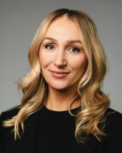 Frau mit welligem, blondem Haar lächelt leicht vor grauem Hintergrund.