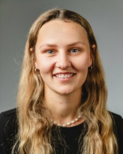 Person mit langen blonden Haaren lächelt vor grauem Hintergrund.