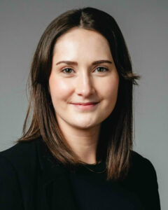 Frau mit schulterlangem Haar trägt schwarzen Blazer vor grauem Hintergrund.