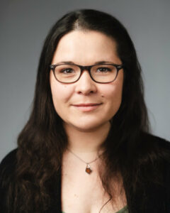 Frau mit Brille und langen Haaren trägt Kette, sitzt vor grauem Hintergrund.