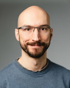 Glatzköpfiger Mann mit Brille und Bart lächelt vor grauem Hintergrund.