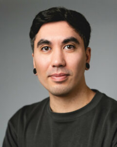 Mann mit schwarzen Ohrsteckern trägt dunkles T-Shirt, neutraler Gesichtsausdruck.