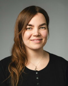 Person mit langen braunen Haaren, trägt ein schwarzes Oberteil, lächelt leicht vor grauem Hintergrund.