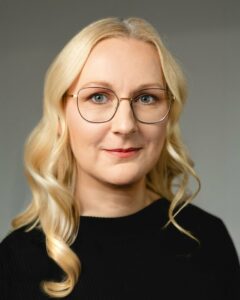 Frau mit runder Brille und lockigem, blondem Haar vor neutralem Hintergrund.