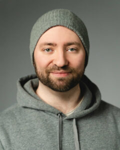 Mann mit grauer Mütze lächelt, trägt grauen Kapuzenpullover vor grauem Hintergrund.