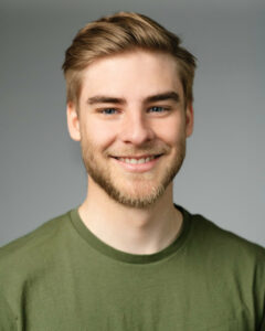 Junger Mann mit Bart und grünem T-Shirt lächelt vor grauem Hintergrund.