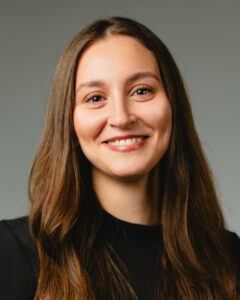 Frau mit langen braunen Haaren lächelt vor grauem Hintergrund.