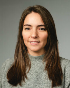 Frau mit langen braunen Haaren trägt grauen Pullover vor grauem Hintergrund.