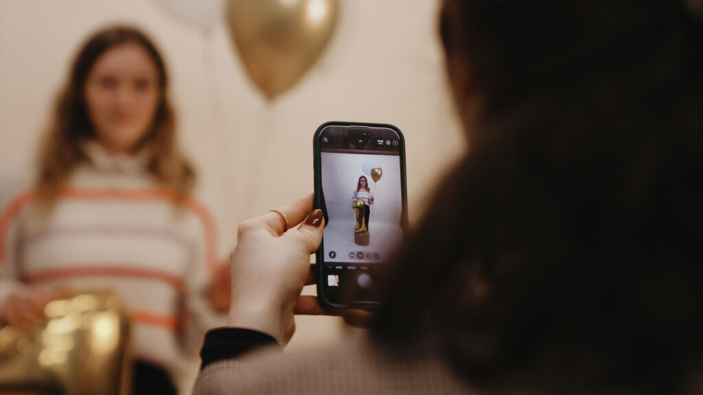 Person hält Smartphone und fotografiert Frau mit goldenem Ballon.