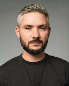 Mann mit kurzem, grauem Haar und Bart, trägt schwarzes T-Shirt vor grauem Hintergrund.